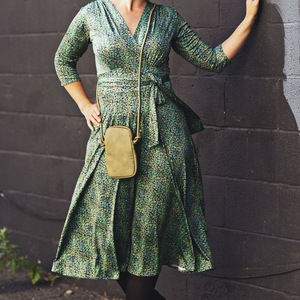 
                  
                    Woman in a green patterned dress standing against a dark brick wall.
                  
                