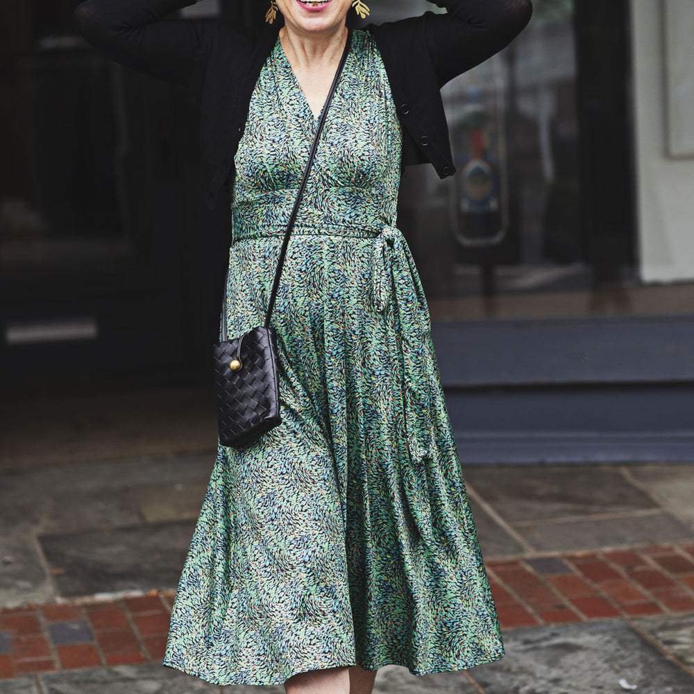 
                  
                    Woman in a green dress with a black cardigan and boots standing on a street.
                  
                